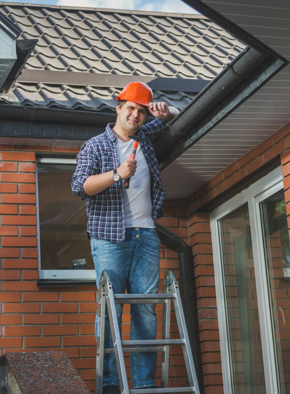 smiling carpenter posing with hammer on step ladder under the roof.jpg